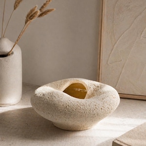 Könnte beinhalten: Eine beige, organisch geformte Keramikschale mit einer großen zentralen Öffnung. Die strukturierte Oberfläche und die neutrale Farbpalette schaffen eine minimalistische Ästhetik. Eine kleinere Vase mit getrockneten Stielen ist im Hintergrund zu sehen.