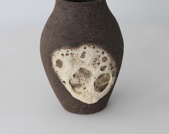 Puede incluir: Un jarrón de cerámica marrón oscuro con una superficie texturizada y un diseño único en forma de corazón blanquecino. El jarrón tiene un cuerpo redondeado y una abertura estrecha, adecuado para exhibir flores o como pieza decorativa.