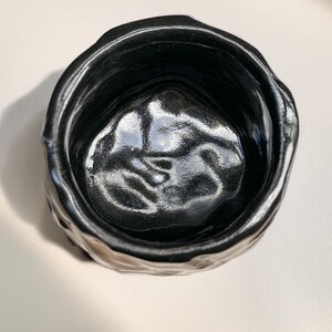 May include: A black ceramic planter with a textured, uneven exterior. The planter has a wide, round opening and a smooth, black interior.