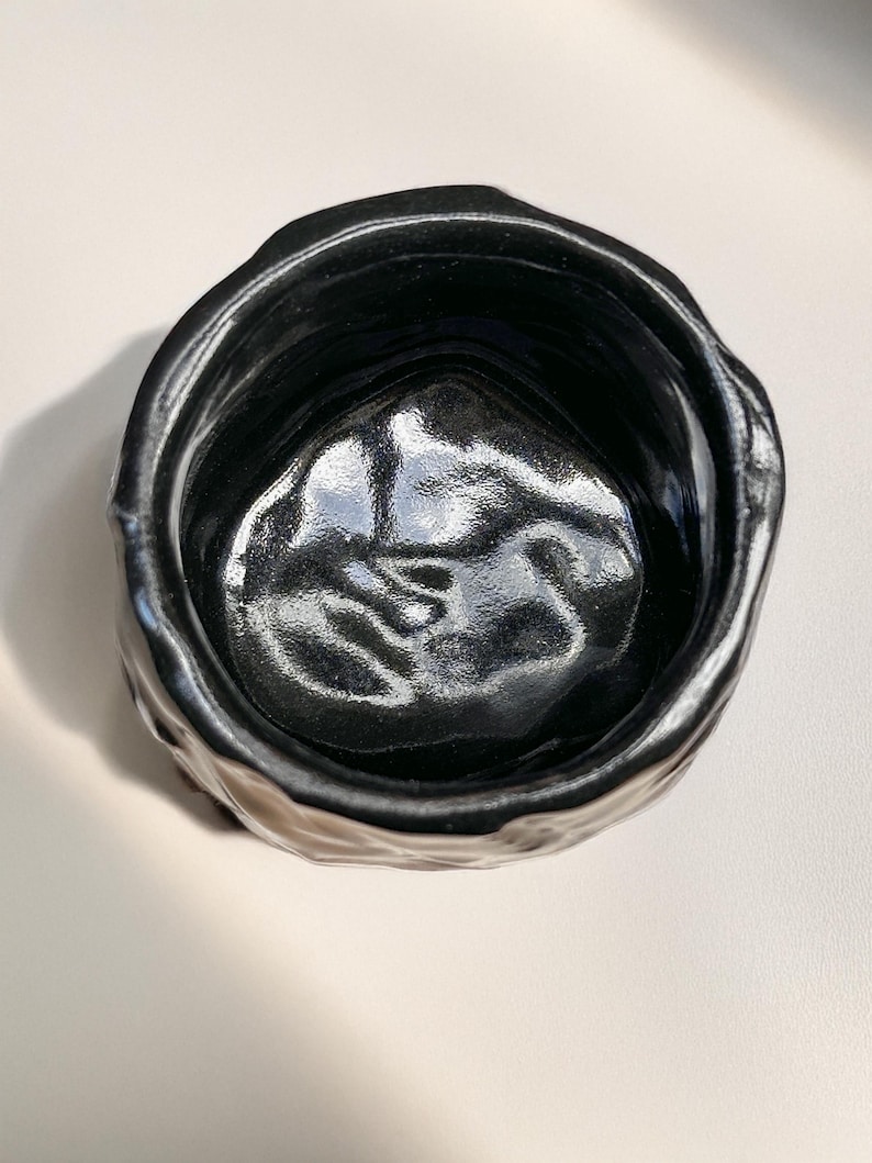 May include: A black ceramic planter with a textured, uneven exterior. The planter has a wide, round opening and a smooth, black interior.