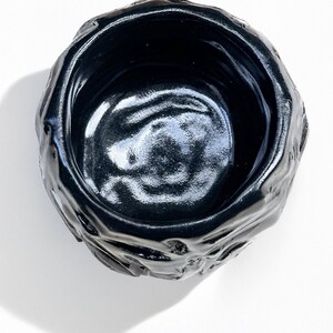 May include: A black ceramic bowl with a rough, textured exterior and a smooth, glossy interior. The bowl is round and has a slightly irregular shape.