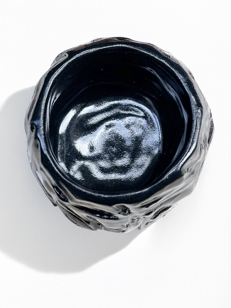 May include: A black ceramic bowl with a rough, textured exterior and a smooth, glossy interior. The bowl is round and has a slightly irregular shape.
