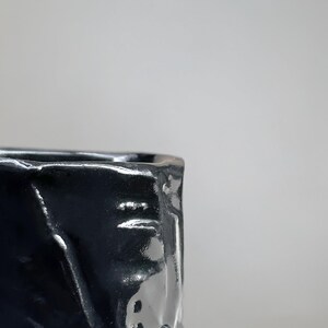 May include: A black ceramic cup with a textured, uneven surface. The cup is glazed and has a glossy finish.
