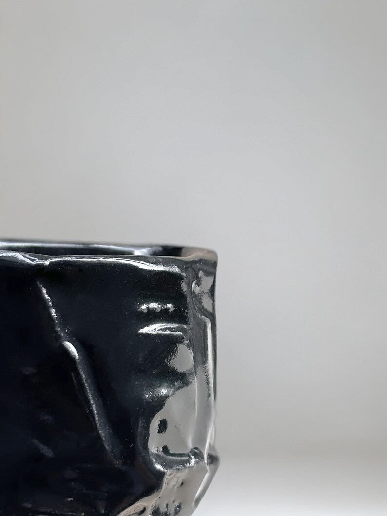May include: A black ceramic cup with a textured, uneven surface. The cup is glazed and has a glossy finish.