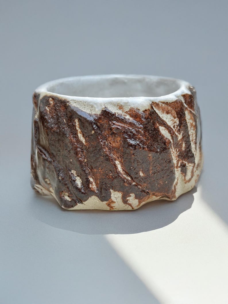 May include: A white ceramic planter with a brown glaze, featuring a textured, rough surface. The planter has a wide, round opening and a slightly rounded base.