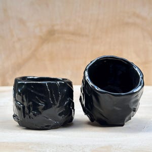 May include: Two black ceramic cups with a textured, rough surface. The cups are empty and have a glossy finish.