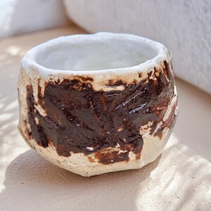 May include: A small, white ceramic cup with brown glaze accents. The cup has a rough, textured surface and an uneven rim.