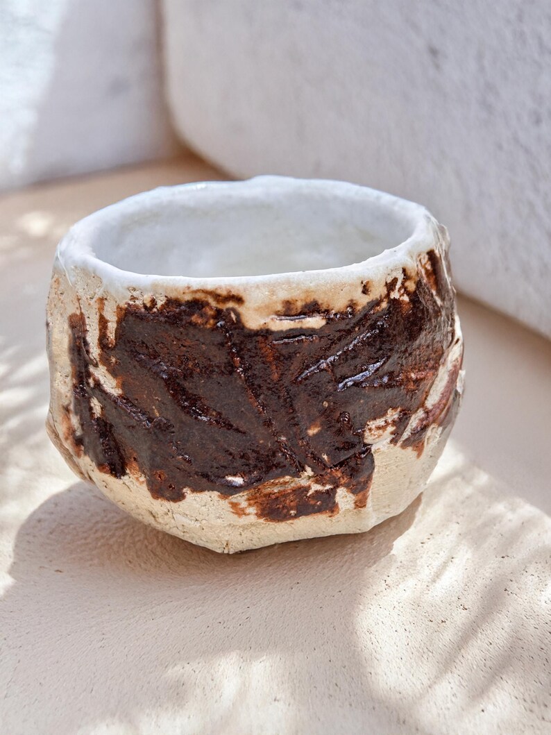 May include: A small, white ceramic cup with brown glaze accents. The cup has a rough, textured surface and an uneven rim.