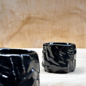 May include: Two black ceramic cups with a textured, rough surface. The cups are glossy and have a dark, almost black, color.