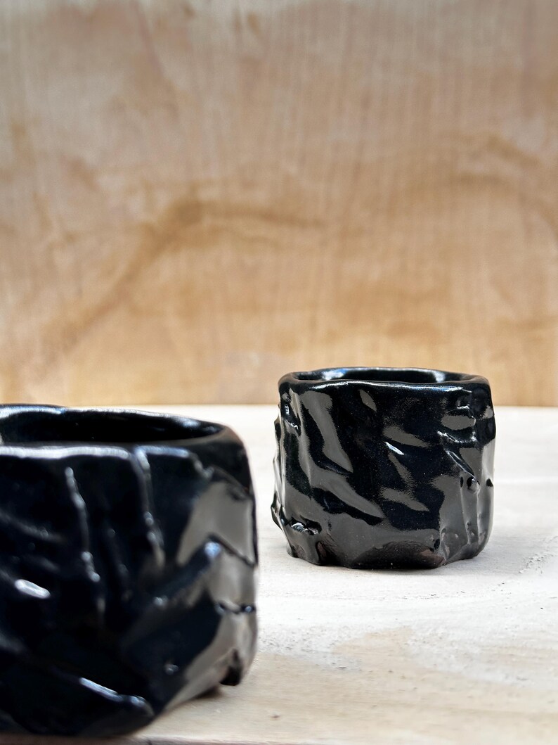 May include: Two black ceramic cups with a textured, rough surface. The cups are glossy and have a dark, almost black, color.