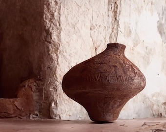 May include: A large, brown, ceramic vase with a textured surface sits on a light brown surface in front of a white wall.