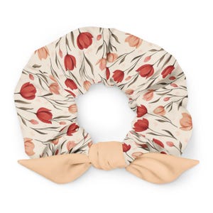 Könnte beinhalten: Ein floraler Haargummi mit cremefarbenem Hintergrund, verziert mit roten und rosa Tulpenillustrationen. Es hat eine pfirsichfarbene Schleife an der Basis. Dieses Haaraccessoire ist perfekt, um jeder Frisur einen Hauch von Frühling zu verleihen.