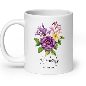May include: White ceramic mug featuring a watercolor floral design with a large purple rose, lavender, pink, and yellow flowers. The name "Kimberly" is written in cursive below the flowers, with "Born in 19XX" underneath.