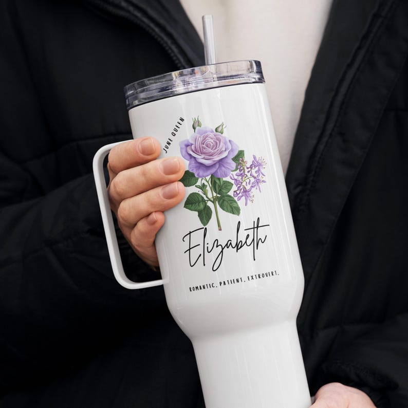 May include: White tumbler with a handle, featuring a purple rose and the name "Elizabeth" in script. The text "June Queen" and personality traits are also included. The tumbler has a clear lid and a straw.