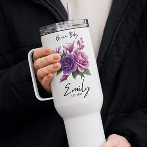 Op de afbeelding: Witte beker met een handvat, versierd met een bloemenontwerp in paarse en roze tinten. De tekst "Gemini Baby" staat bovenaan, met de naam "Emily" en "EST 1995" onder de bloemen. Inclusief een doorzichtig deksel en een rietje.