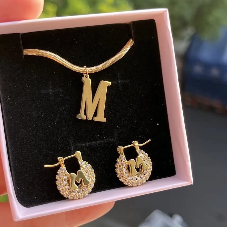 May include: A gold necklace and earrings set featuring the letter "M" with clear crystal accents. The necklace has a delicate chain and the earrings are hoop style.