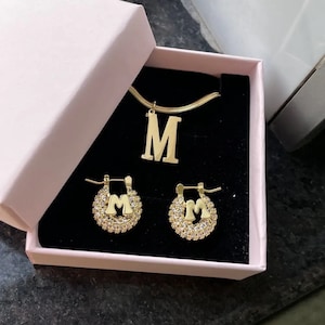 May include: A gold necklace and earrings set featuring the letter "M" with sparkling rhinestones. The jewelry is displayed in a pink box.