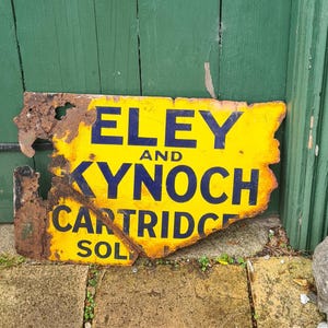 May include: A weathered yellow sign with the text "ELEY AND KYNOCH CARTRIDGE SOLD" in black lettering. The sign is damaged and rusty, and is mounted on a green wooden door.