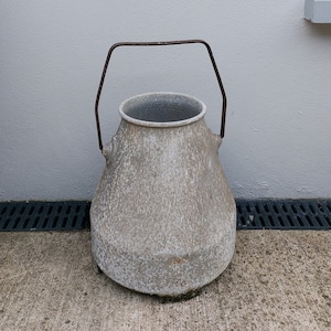 Puede incluir: Un contenedor grande de metal gris con un asa de metal oxidado. El contenedor está sobre una superficie de hormigón.