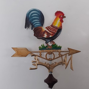 Op de afbeelding: Een gouden metalen windwijzer met een geschilderde haan, pijl en kompaspunten. De haan is rood, geel en bruin met een blauwe staart. De pijl wijst naar het oosten.