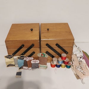 Puede incluir: Dos cajas de costura de madera con bisagras negras y un pomo negro en la parte superior. Las cajas están abiertas y llenas de carretes de hilo, botones y otros materiales de costura. También hay varias etiquetas para diferentes tipos de hilo.