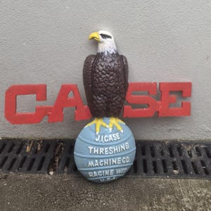 May include: A cast metal sign featuring a brown and white eagle perched on a light blue globe. The globe displays the text J.I.CASE THRESHING MACHINE CO. RACINE WIS. U.S.A. Behind the eagle is a large, bold, red block letter sign reading CASE.