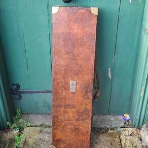 Op de afbeelding: Een rechthoekige bruin lederen koffer met messing hoekbeschermers en een metalen plaat. De koffer staat tegen een verweerde groene deur met zwart beslag. De koffer lijkt een antiek stuk te zijn.