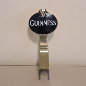 Puede incluir: Un grifo de cerveza Guinness con un letrero ovalado negro con el logotipo de Guinness en letras blancas. El grifo es de color dorado y tiene una base curvada. La parte superior del letrero tiene un s&iacute;mbolo de arpa dorada.