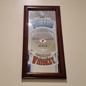 Puede incluir: Espejo rectangular con marco de madera marr&oacute;n oscuro. El espejo muestra la palabra "POWERS" en azul y "WHISKEY" en rojo. El centro tiene un logotipo con la letra "P".