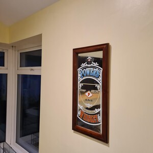 Puede incluir: Espejo rectangular con marco de madera marr&oacute;n oscuro. El espejo presenta un anuncio vintage de "Powers Whiskey" con texto en azul, blanco y rojo. El espejo est&aacute; colgado en una pared de color crema.