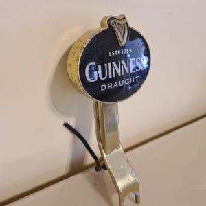 May include: A vintage Guinness draught tap handle. The handle is gold-colored with a black and white circular emblem featuring the Guinness logo and the text "DRAUGHT". The emblem also includes a harp symbol and the text "ESTD 1759".