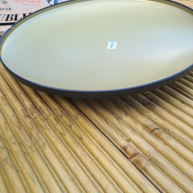 May include: A large, round serving tray with a gold-colored interior and a dark rim. The tray is set on a wooden surface with a natural finish. A small white label is visible on the tray's surface.