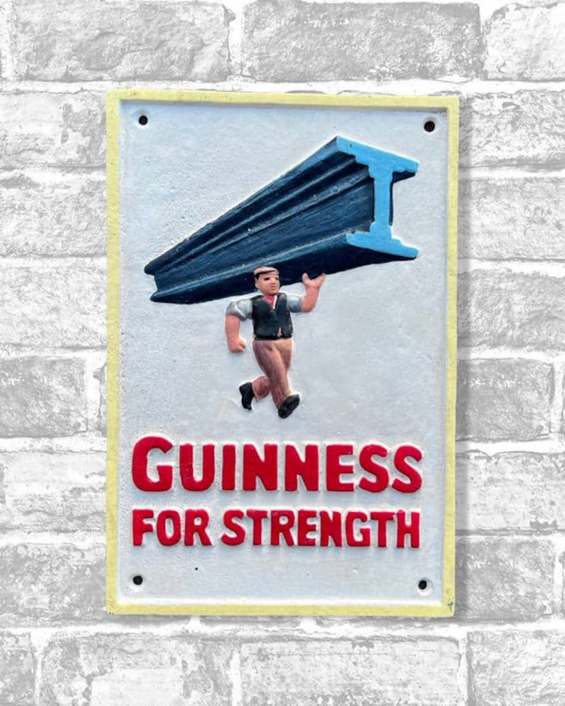 May include: A rectangular sign with a yellow border and the words "GUINNESS FOR STRENGTH" in red. The sign features a figure carrying a blue I-beam. The background is a light gray color, and the sign is mounted on a brick wall.