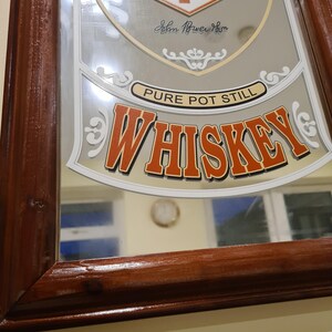 Puede incluir: Un espejo rectangular con marco de madera con la palabra "WHISKEY" en letras naranjas. El espejo tambi&eacute;n tiene las palabras "PURE POT STILL" y una firma. El marco es de color marr&oacute;n oscuro.