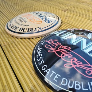 May include: Two oval Guinness signs. One is cream-colored with black and orange text, including "Guinness" and "Gate Dublin." The other is black with white and pink text, including "Arthur Guinness" and "St. James's Gate Dublin."