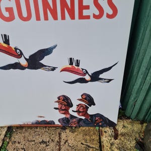 May include: Vintage Guinness advertisement featuring toucans and two men in military-style hats. The word "GUINNESS" is printed in large red letters at the top. The toucans have red, yellow, and black beaks and are in flight. The men have mustaches and are looking up.