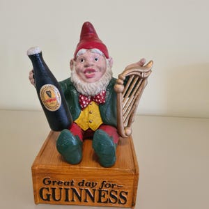 May include: A ceramic gnome figurine holding a Guinness bottle and a harp. The gnome wears a red hat, green jacket, yellow vest, and red bow tie. The base reads "Great day for Guinness."