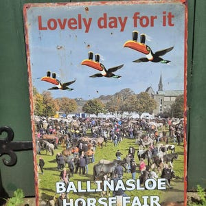 May include: A vintage metal sign with the text "Lovely day for it" and images of toucans carrying pints of beer. The sign also features the text "Ballinasloe Horse Fair" and a crowd of people and horses.