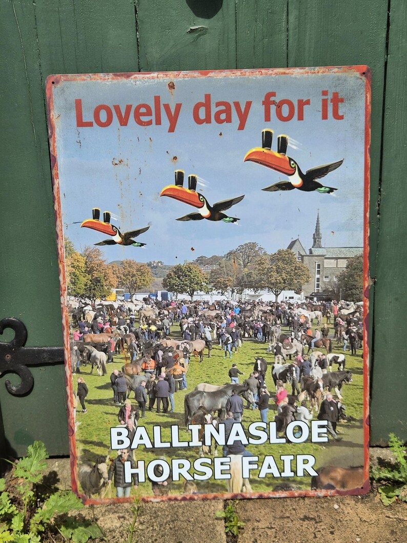 May include: A vintage metal sign with the text "Lovely day for it" and images of toucans carrying pints of beer. The sign also features the text "Ballinasloe Horse Fair" and a crowd of people and horses.