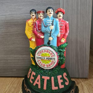 May include: A black cast iron doorstop featuring the Beatles in their Sgt. Pepper's Lonely Hearts Club Band attire. The doorstop has a green base with the band's name in pink letters.