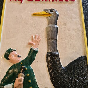 K&ouml;nnte beinhalten: Ein Vintage-Schild aus Gusseisen mit gelbem Rand und dem Text "My Goodness My GUINNESS" in Rot. Das Schild zeigt eine Cartoon-Illustration eines Mannes in gr&uuml;ner Uniform, der eine Flasche Bier h&auml;lt, und eine Strau&szlig; mit langem Hals.