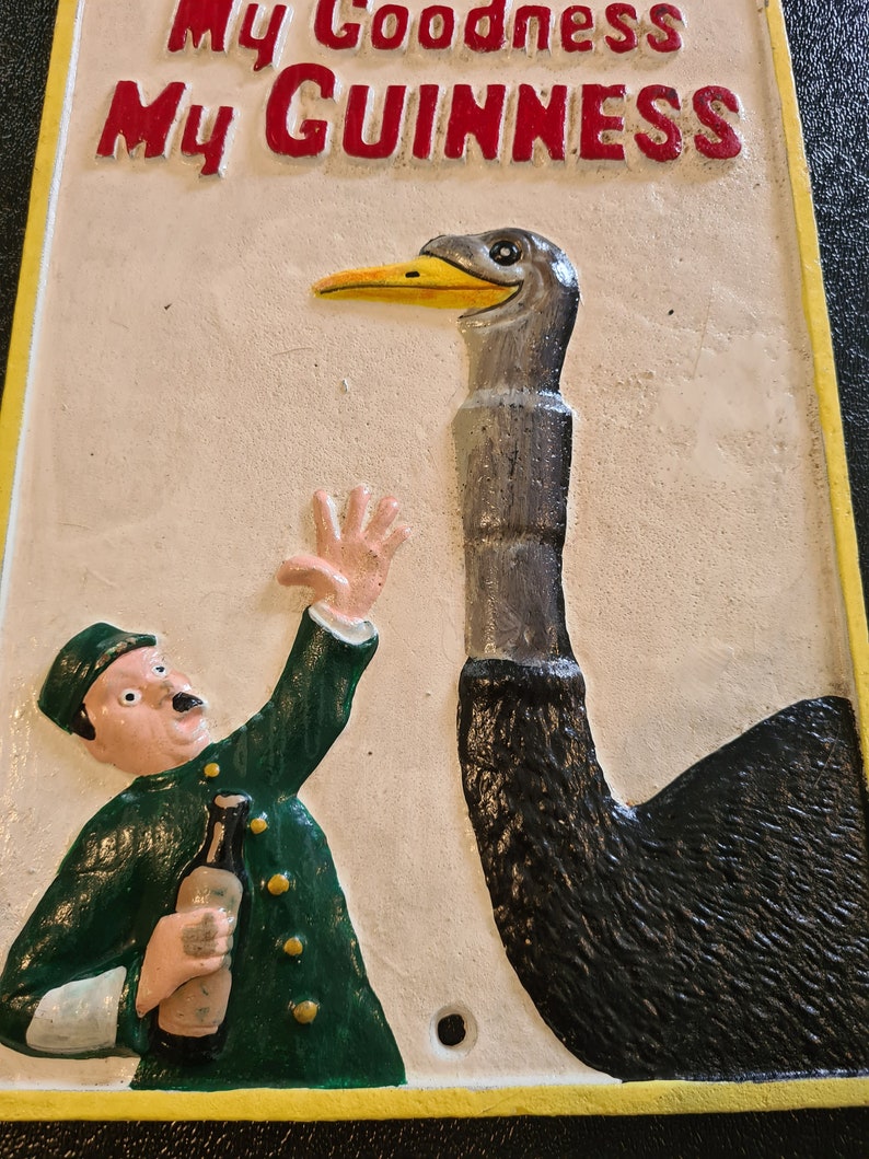 K&ouml;nnte beinhalten: Ein Vintage-Schild aus Gusseisen mit gelbem Rand und dem Text "My Goodness My GUINNESS" in Rot. Das Schild zeigt eine Cartoon-Illustration eines Mannes in gr&uuml;ner Uniform, der eine Flasche Bier h&auml;lt, und eine Strau&szlig; mit langem Hals.