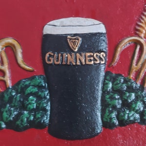 May include: A red sign with the word "IRELAND" at the top and "GUINNESS" in gold lettering on a black pint glass. The sign has green foliage and gold accents, with the word "INN" at the bottom.