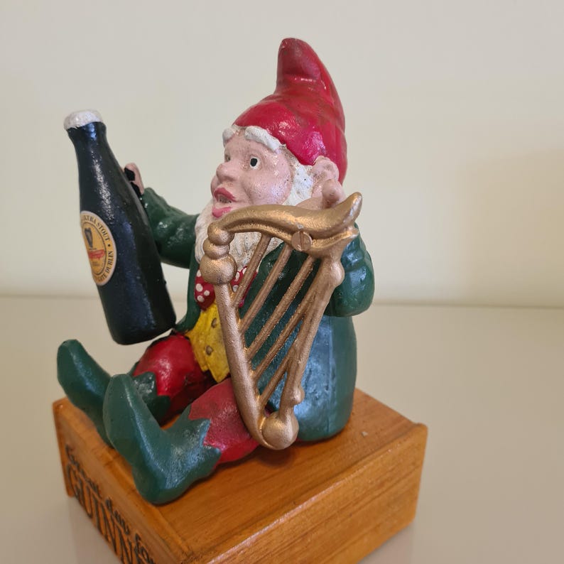 May include: A whimsical figurine of a gnome holding a stout bottle and a harp. The gnome wears a red hat, green jacket, and red pants. The base is a wooden block with text. The bottle is black with a gold label.