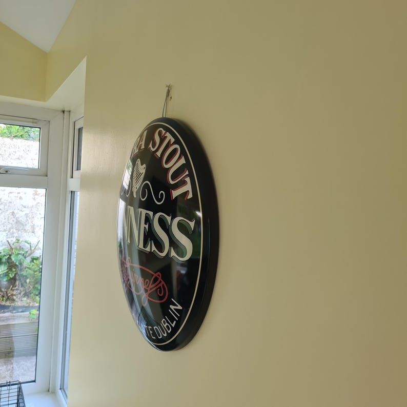 May include: A black and white Guinness Stout sign with a glossy finish. The oval sign features the Guinness logo and the words "Dublin" and "Stout". The sign is hanging on a light yellow wall.
