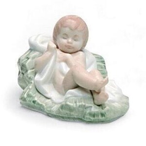 Può includere: Una statuetta in porcellana di un bambino Gesù sdraiato su un letto di foglie verdi, avvolto in un panno bianco. Il bambino ha gli occhi chiusi e un'espressione pacifica.