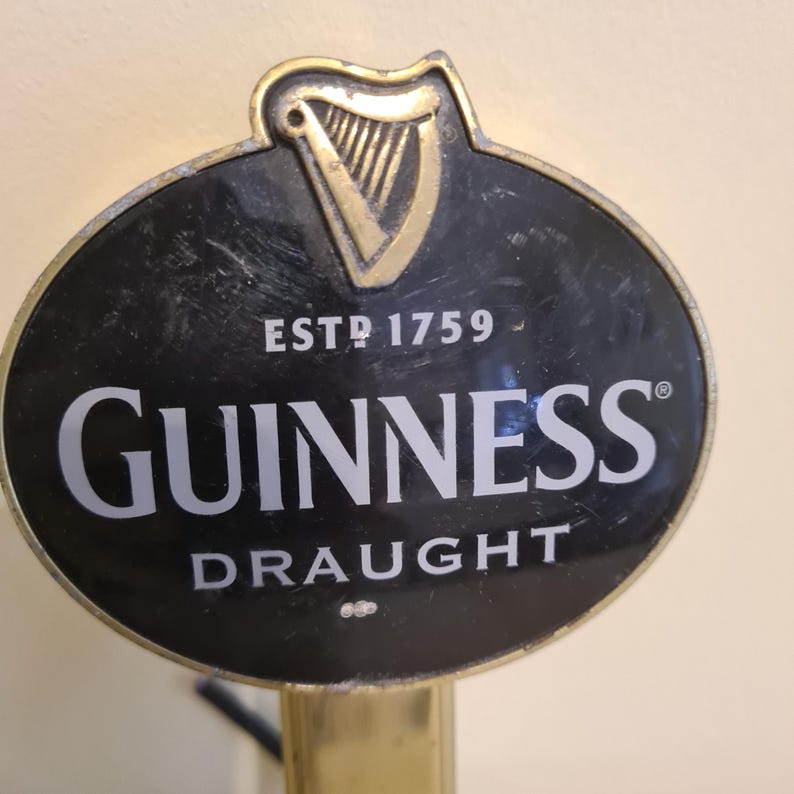 May include: A black and gold Guinness draught tap handle. The oval-shaped sign features the Guinness logo in white, the word "DRAUGHT" below, and "ESTD 1759" above. A gold harp is at the top.
