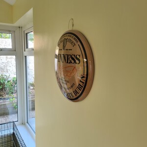 May include: A round Guinness sign with a black border and a light brown background. The sign features the Guinness logo and text, including "Dublin" and "Stout". The sign is hanging on a yellow wall.