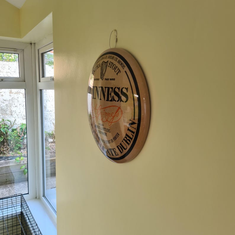 May include: A round Guinness sign with a black border and a light brown background. The sign features the Guinness logo and text, including "Dublin" and "Stout". The sign is hanging on a yellow wall.