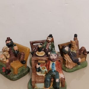 May include: Four ceramic figurines of men sitting on park benches. Each figurine is wearing different clothing and has a different expression. One figurine is holding a beer, another is holding a cup, and another is holding a cat. The figurines are painted in realistic colors and details.